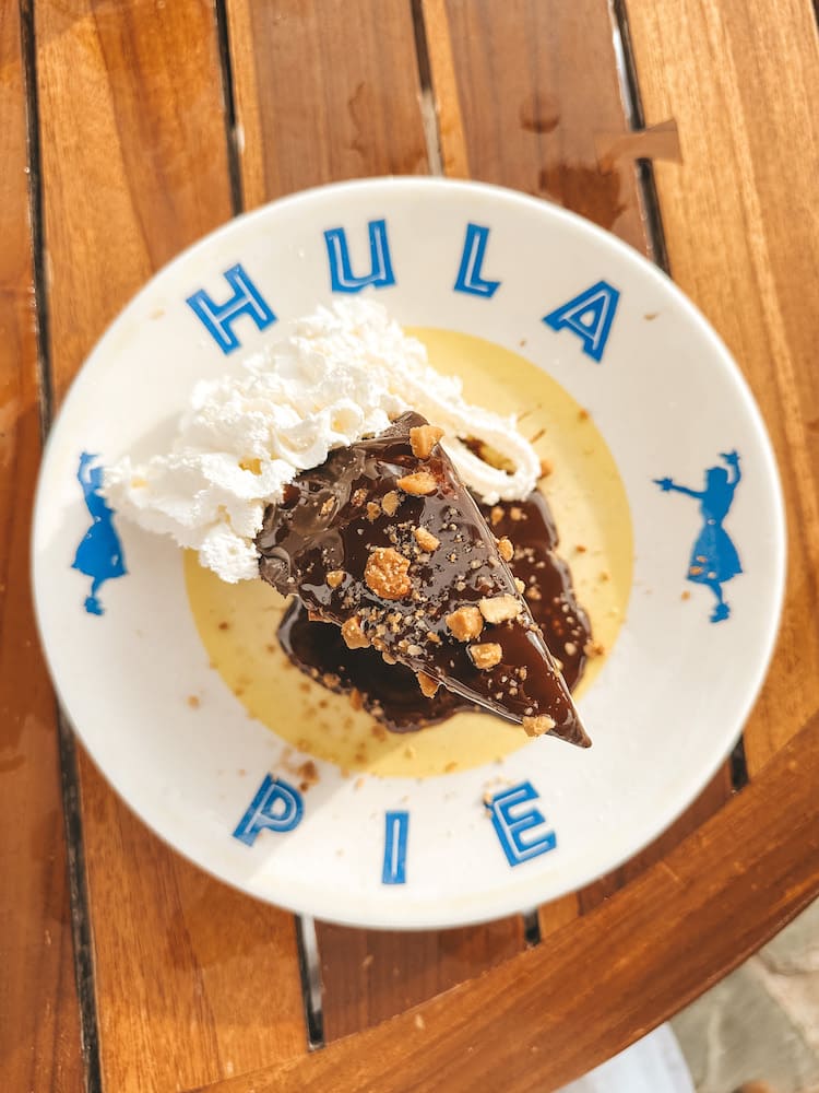 A slice of Hula Pie on a plate that says, "Hula Pie," sitting on a wooden table at Duke's Maui.