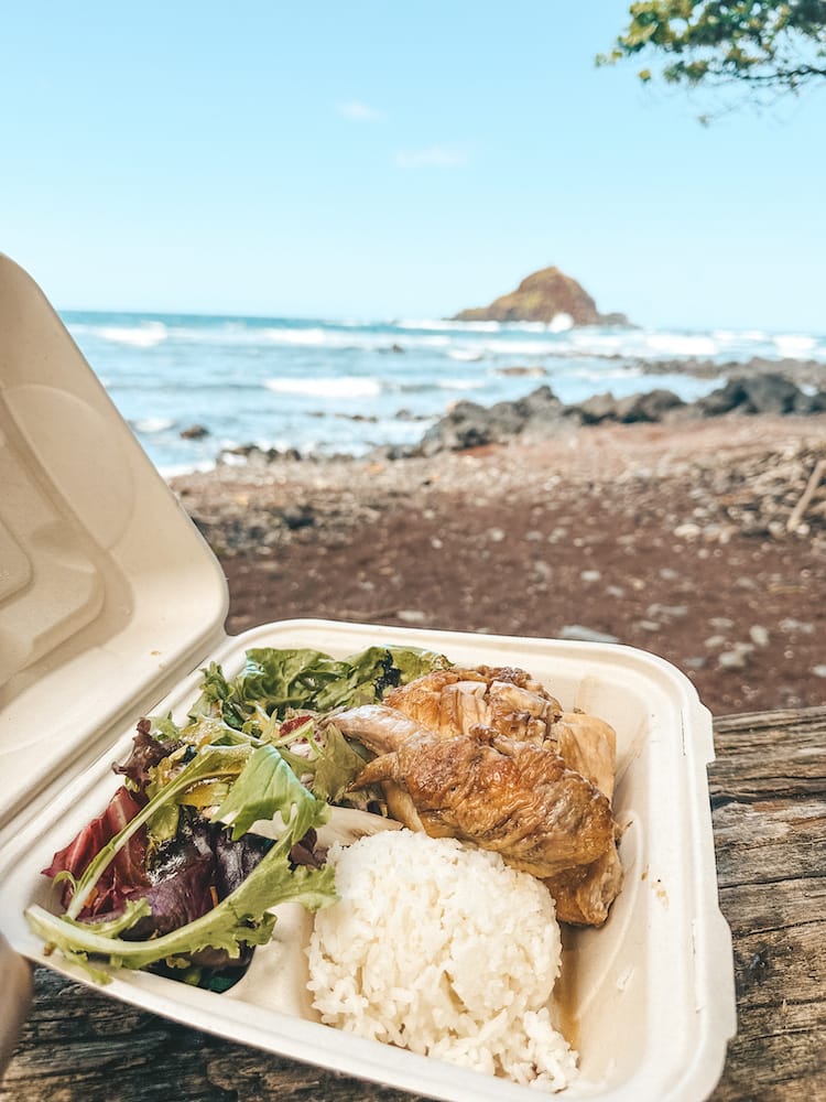 A disposable carton containing huli huli chicken, white rice, and a green salad in front of a red sand beach and the ocean on the Road to Hana in Maui.