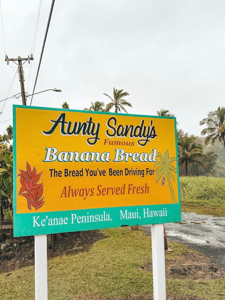 A yellow and green sign that says "Aunt Sandy's Famous Banana Bread. The Bread You've Been Driving For. Always Served Fresh. Ke'Anae Peninsula, Maui, Hawaii"