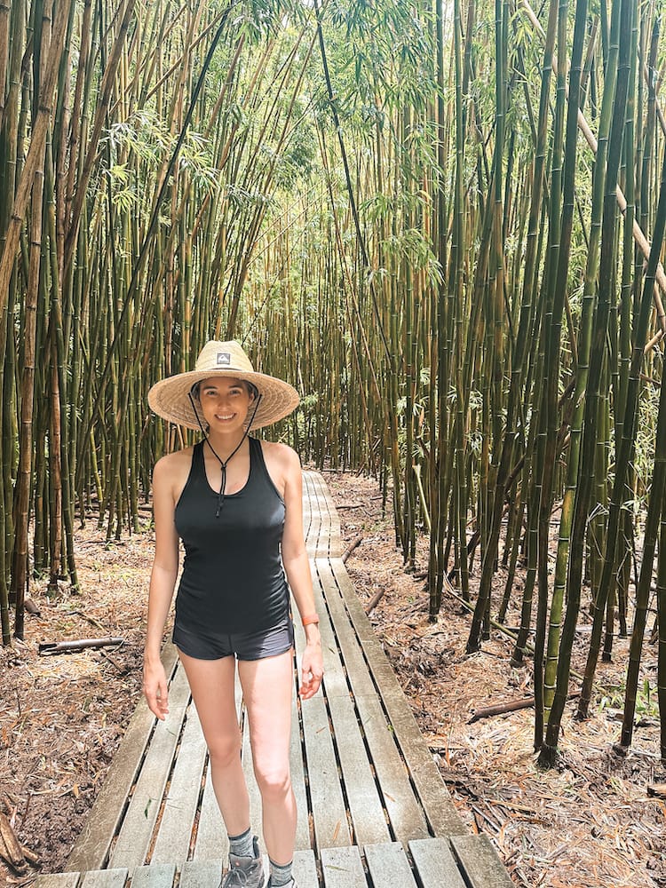 A woman in a black tank top, shorts, and a straw hat standing on a wooden platform in the middle of a bamboo forest