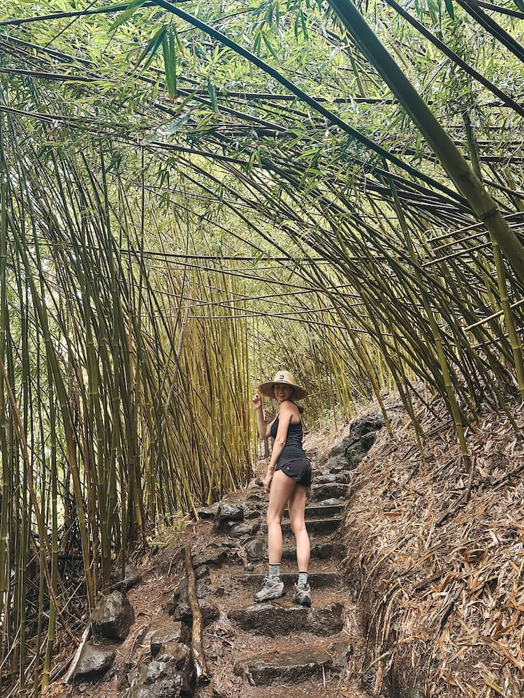 A woman in a black tank top, shorts, and straw hat standing on rocky stairs in the middle of a bamboo forest