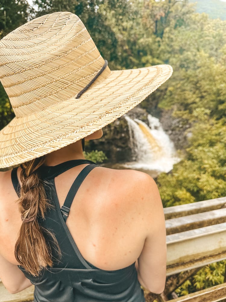 A woman wearing a black tank top, a straw hat, with braided hair looking at a waterfall over a bridge.