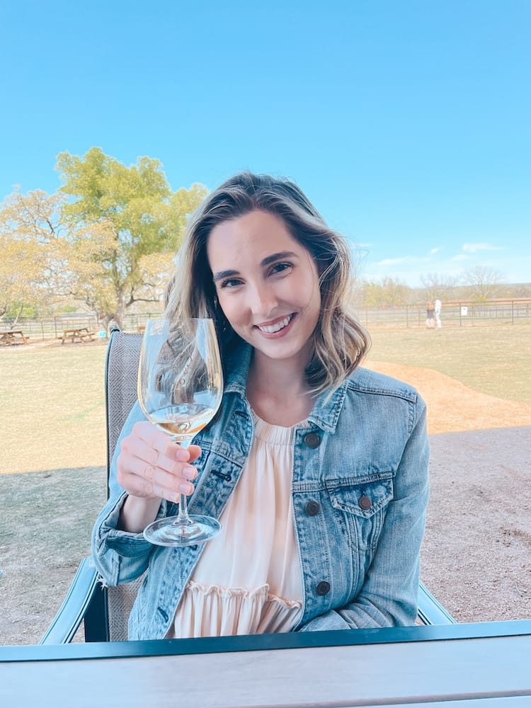 A woman wearing a white dress and denim jacket holding a glass of white wine in the Texas Hill Country