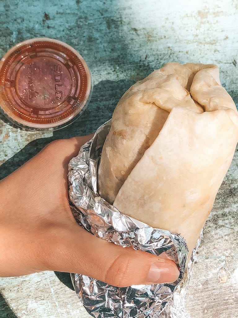 A breakfast burrito from Kono's Northshore wrapped in tin foil with a red salsa sitting on a blue picnic bench.