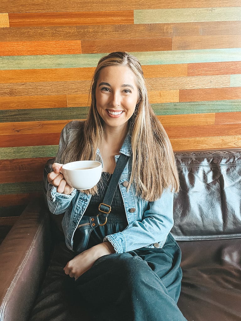 A woman in a black jumpsuit and denim jacket drinking a cup of coffee in Storyville Coffee in Seattle.