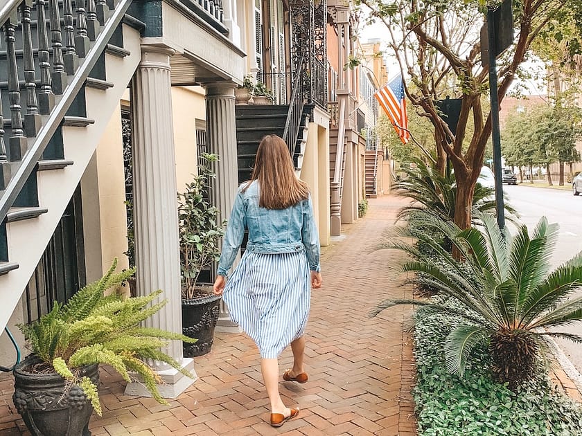 A girl with medium-length brown hair with a blue dress walking down a cobblestone street surrounded by palm trees.