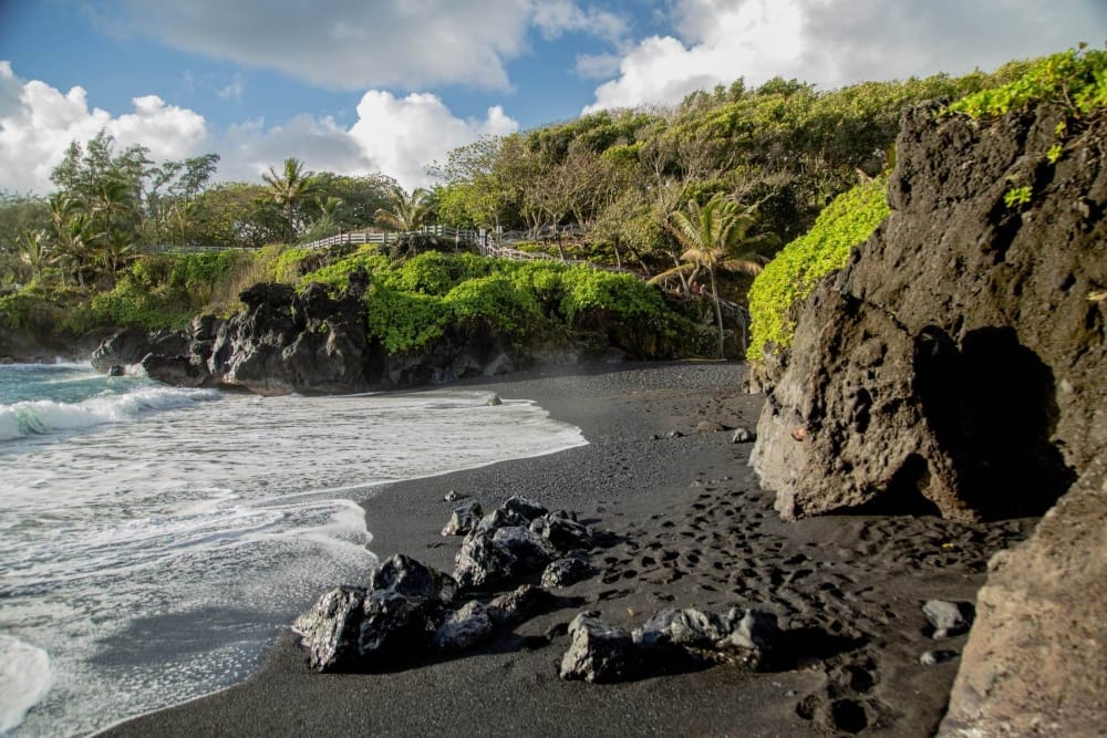 A black sand beach in Maui, surrounded by green vegetation and blue waves lapping up on the shore.