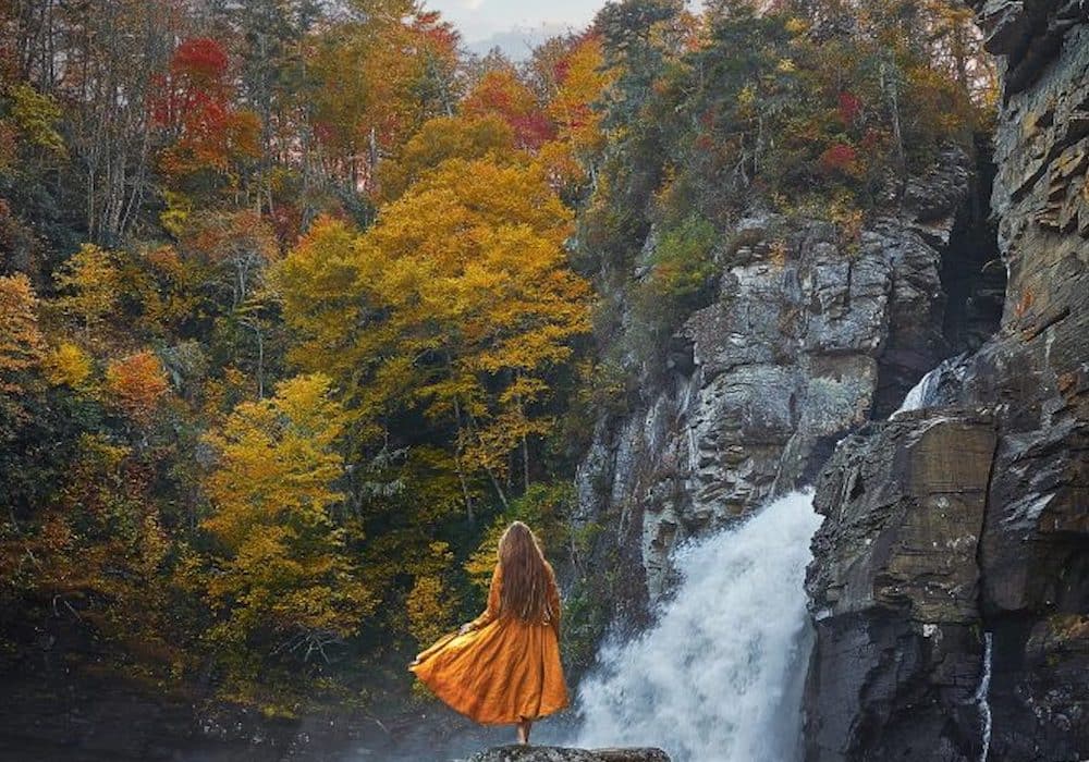 A woman standing in an orange dress in front of yellow and orange autumn leaves and a waterfall flowing down a rock in Asheville, North Carolina.