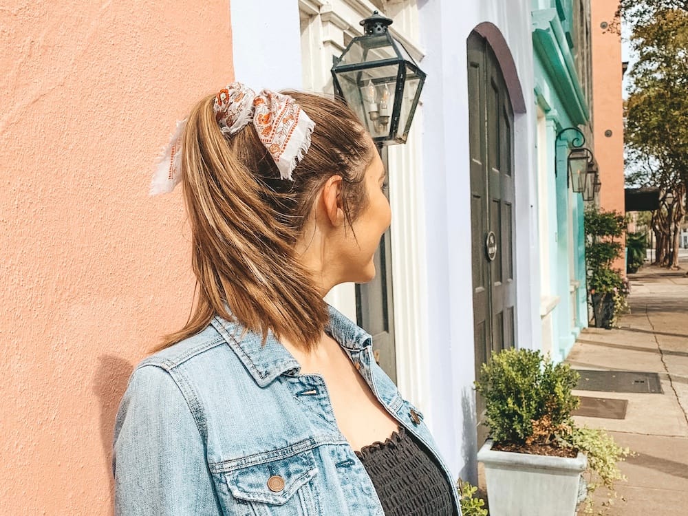A woman wearing a black top, denim jacket, and ponytail standing in front of colorful painted houses on Rainbow Row in Charleston.