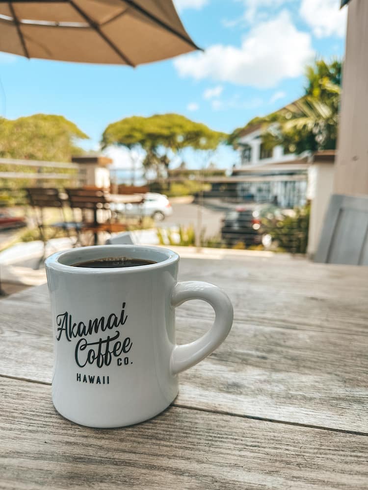 A white coffee mug that says Akamai Coffee Co. Hawaii sitting on a wooden table with tropical trees and a parking lot in the background