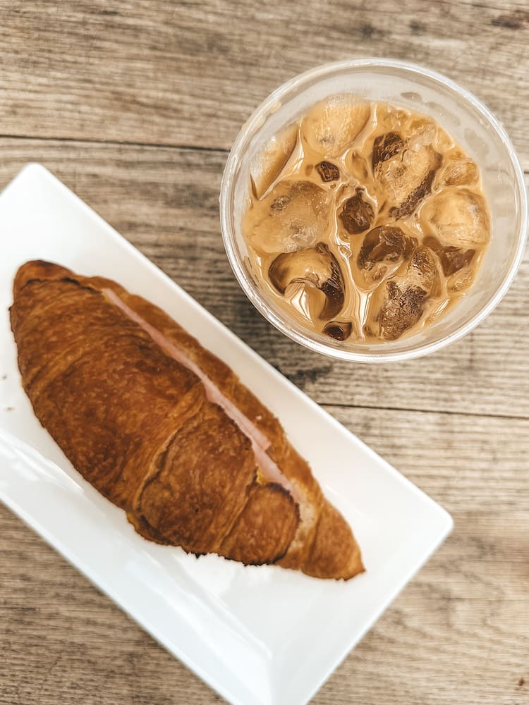 An iced coffee and a croissant sandwich with ham sitting on a wooden table.