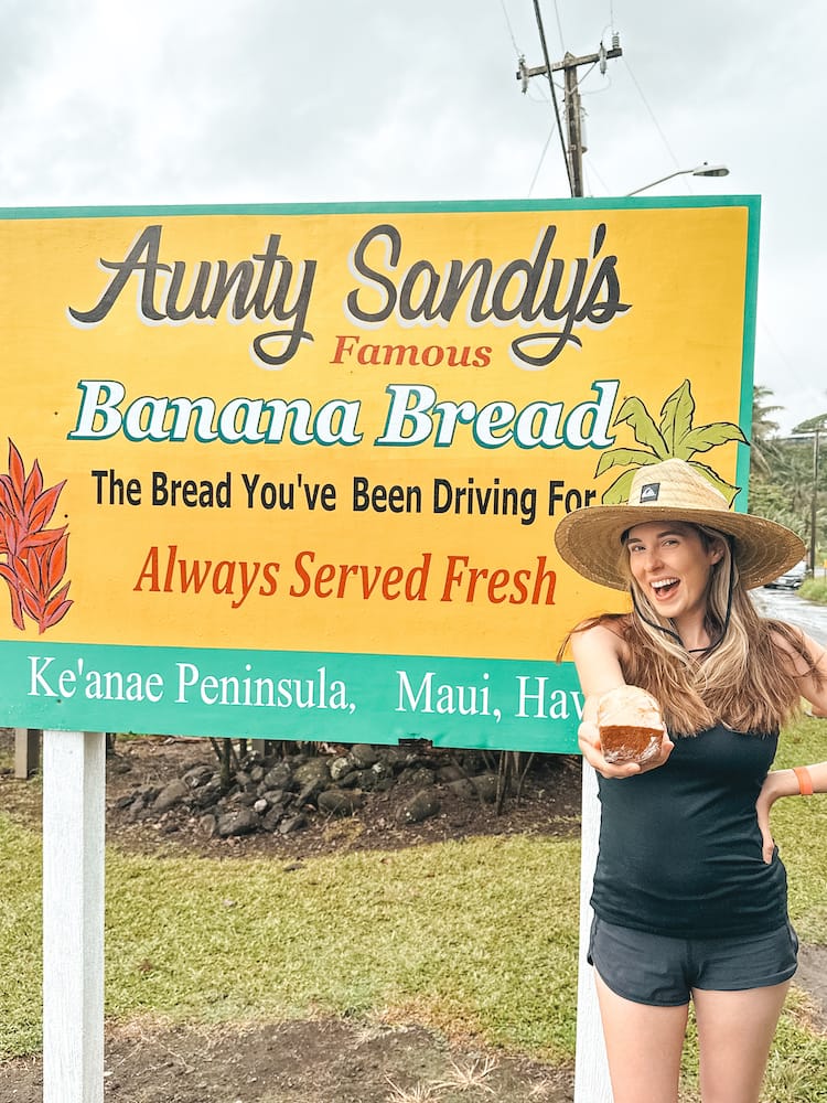 A woman in black athleticwear and a straw hat holding a loaf of banana bread out in front of a yellow and green sign that reads: "Aunt Sandy Famous Banana Bread. The Bread You've Been Driving For, Always Served Fresh." One of the best places to eat in Maui on the Ke'anae Peninsula