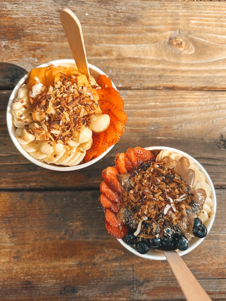 Two acai bowls on a wooden table. One is filled with bananas, blueberries, and strawberries, and the other is filled with bananas, strawberries, and papaya.