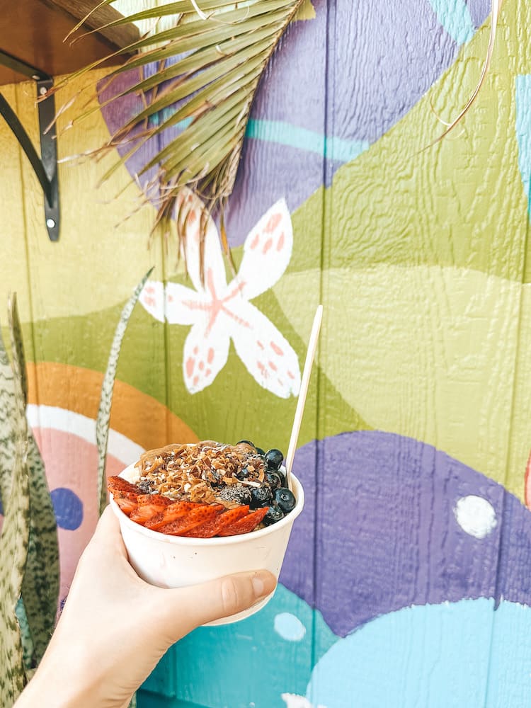An acai bowl topped with blueberries, strawberries, nut butters, chia seeds, and other toppings in front of a colorful mural.