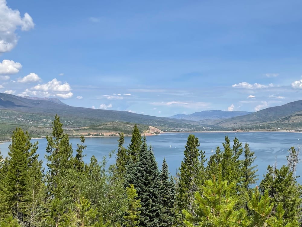 Several green trees lining the perimeter of a blue lake in Colorado with some mountains in the background.