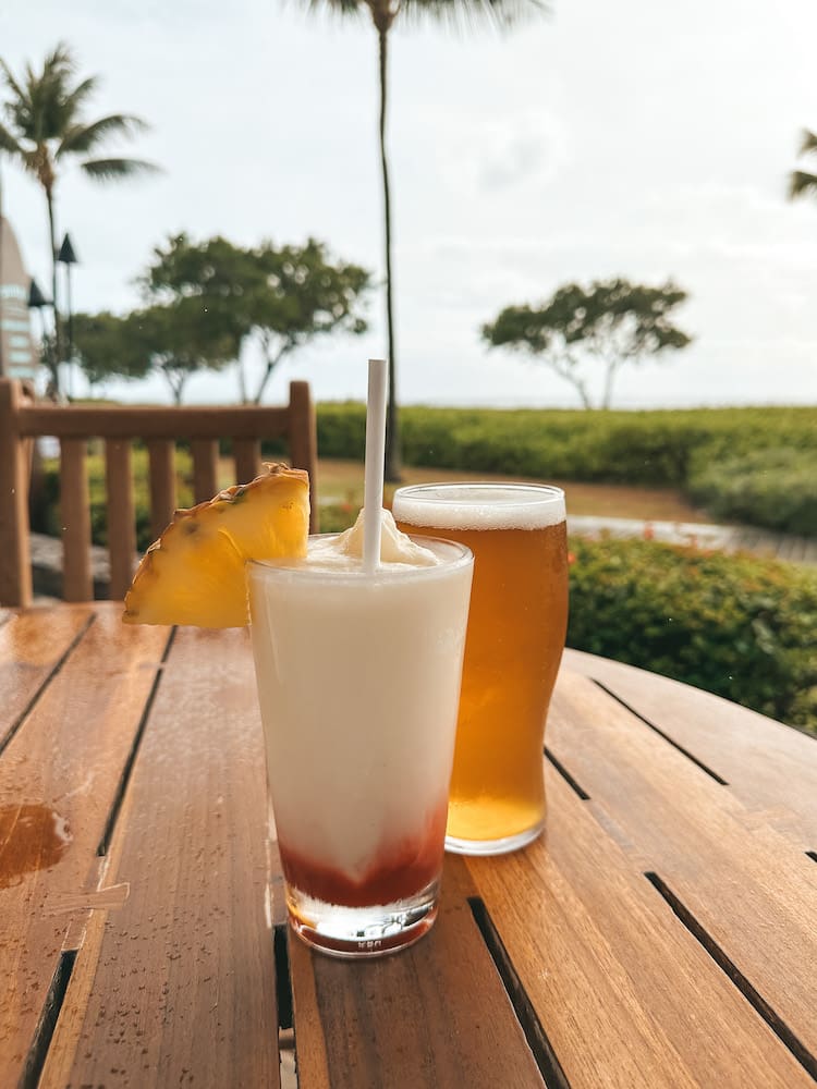 A beer and a pina colada with strawberry flavor sitting on a table overlooking the ocean in Maui.