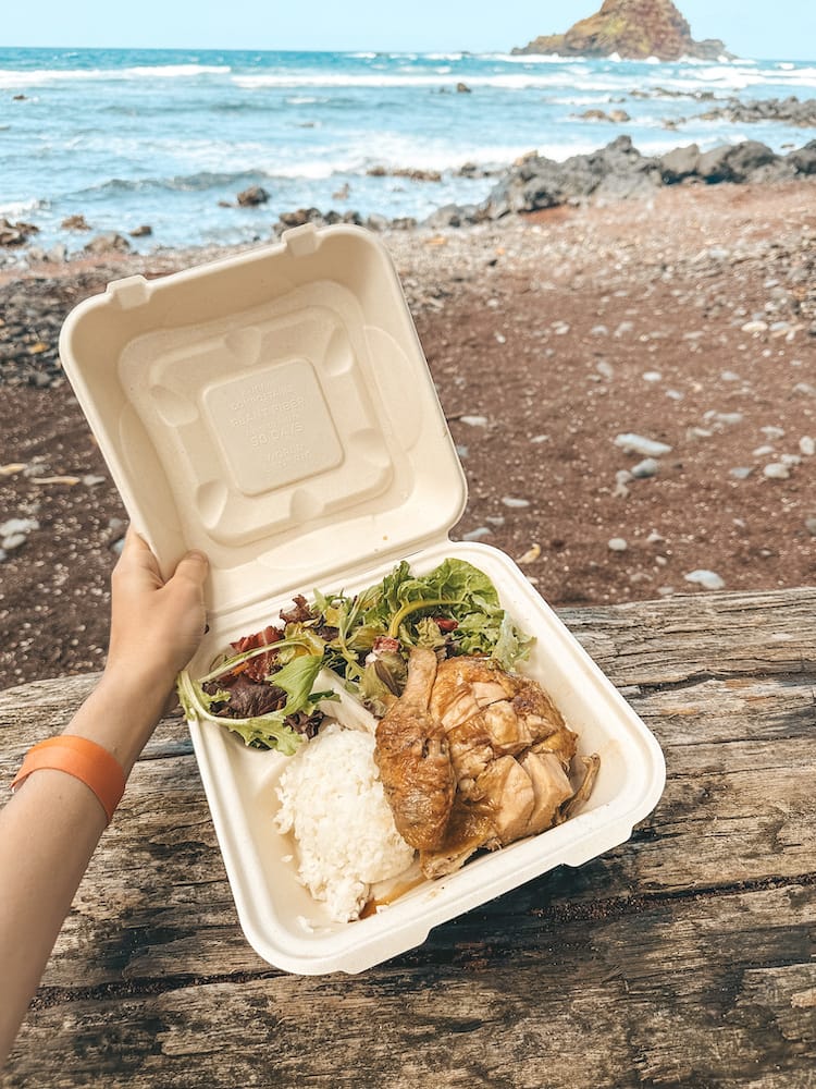 A cardboard takeout box filled with chicken, ride, and a salad on a red sand beach in Maui