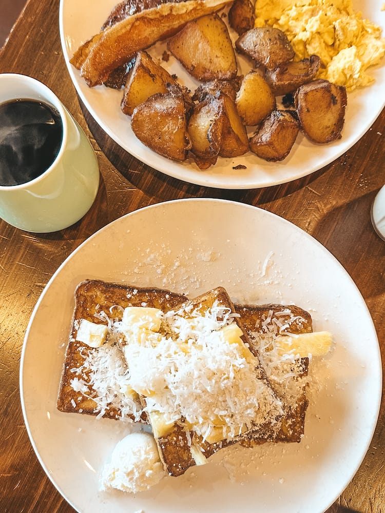 A breakfast spread with French toast topped with pineapple and coconut, hot coffee in a mug, and a plate with eggs, bacon, and potatoes in the background.