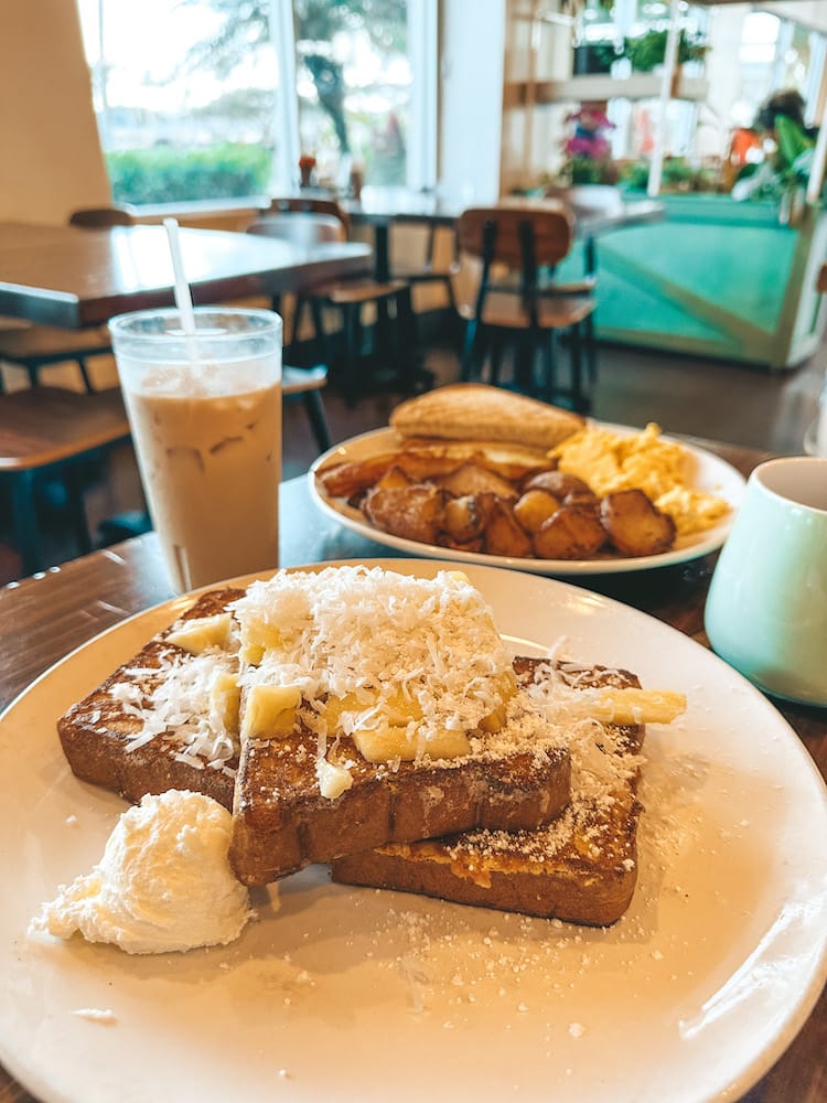 A breakfast spread with French toast topped with pineapple and coconut, iced coffee, and a plate with eggs, toast, and potatoes in the background.