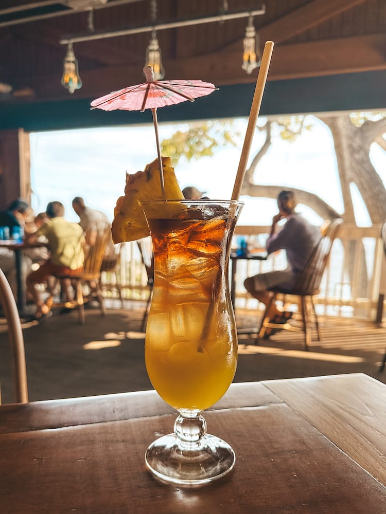 A Mai Tai cocktail in a tall glass with a pineapple and pink umbrella decorating it sitting on a table at one of the best restaurants in Maui.