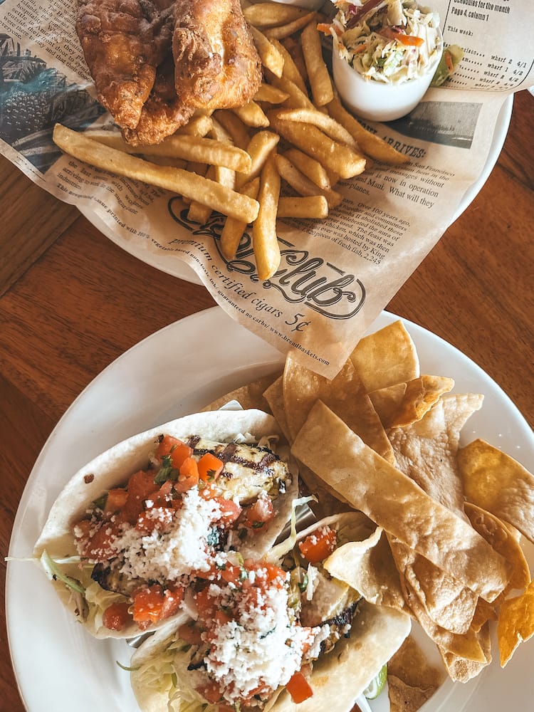 Two plates sitting on a wooden table. One include fish tacos and chips, and the other includes fish and chips.