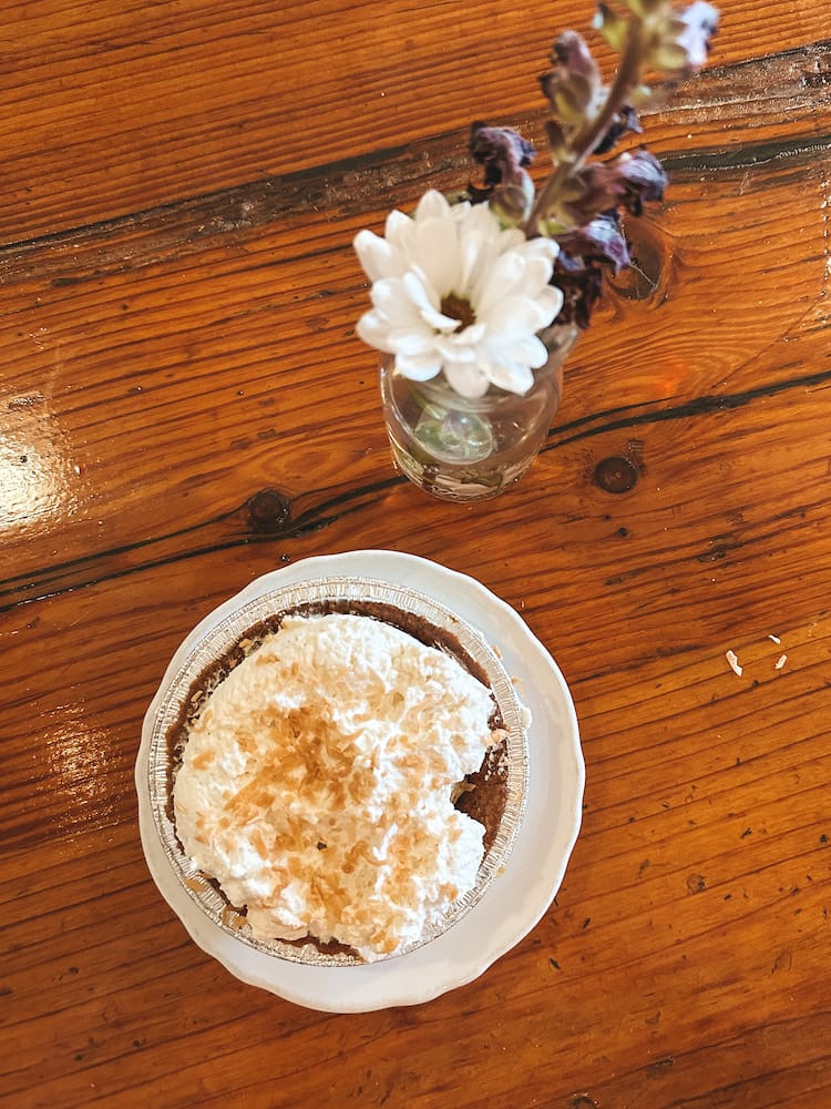 A small coconut cream pie sitting on a wooden table with a small vase holding white and purple flowers.