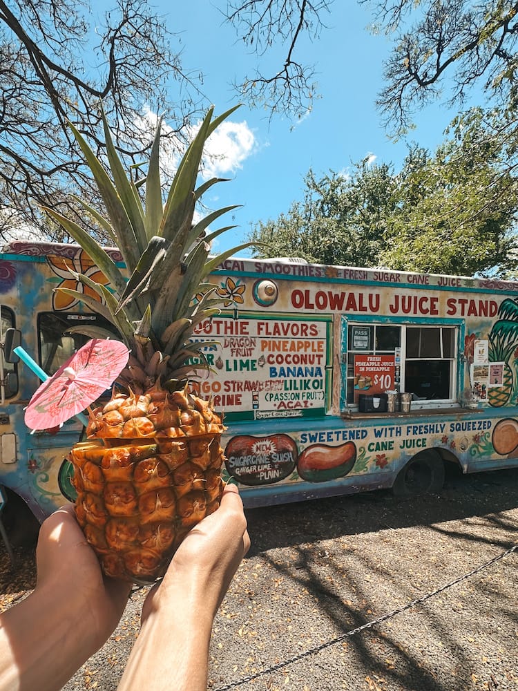 A drink in a pineapple with a pink umbrella in front of a colorful food truck that reads "Olowalu Juice Stand"