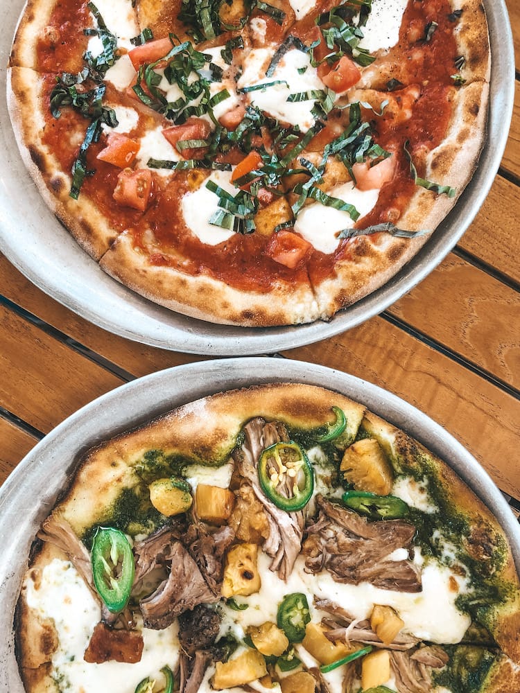 Two pizzas sitting on a wooden table at Monkeypod Kitchen. One is a margarita pizza, and one contains kalua pork, pineapple, and jalapenos.