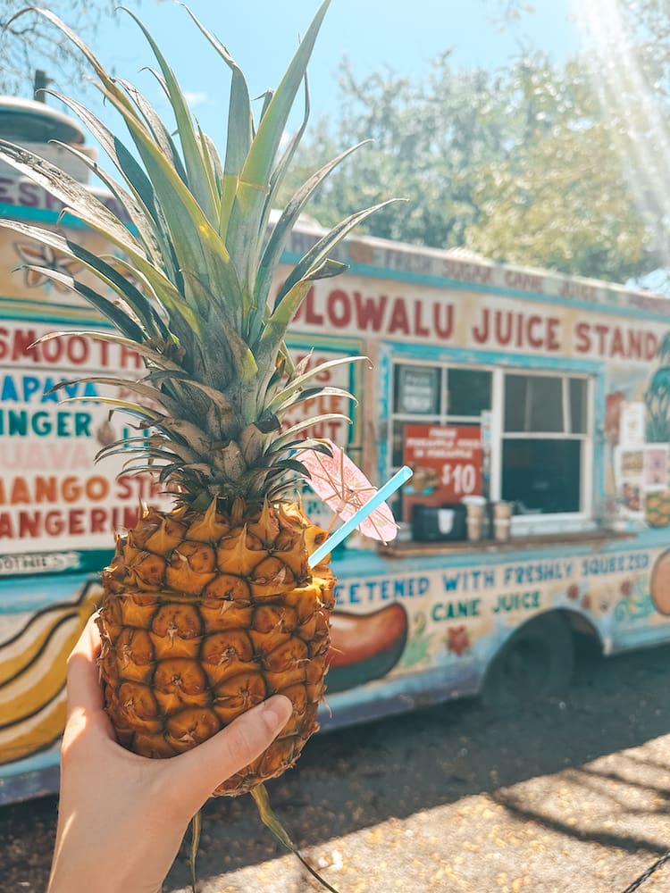 A drink in a pineapple with a pink umbrella in front of a colorful food truck that reads "Olowalu Juice Stand"