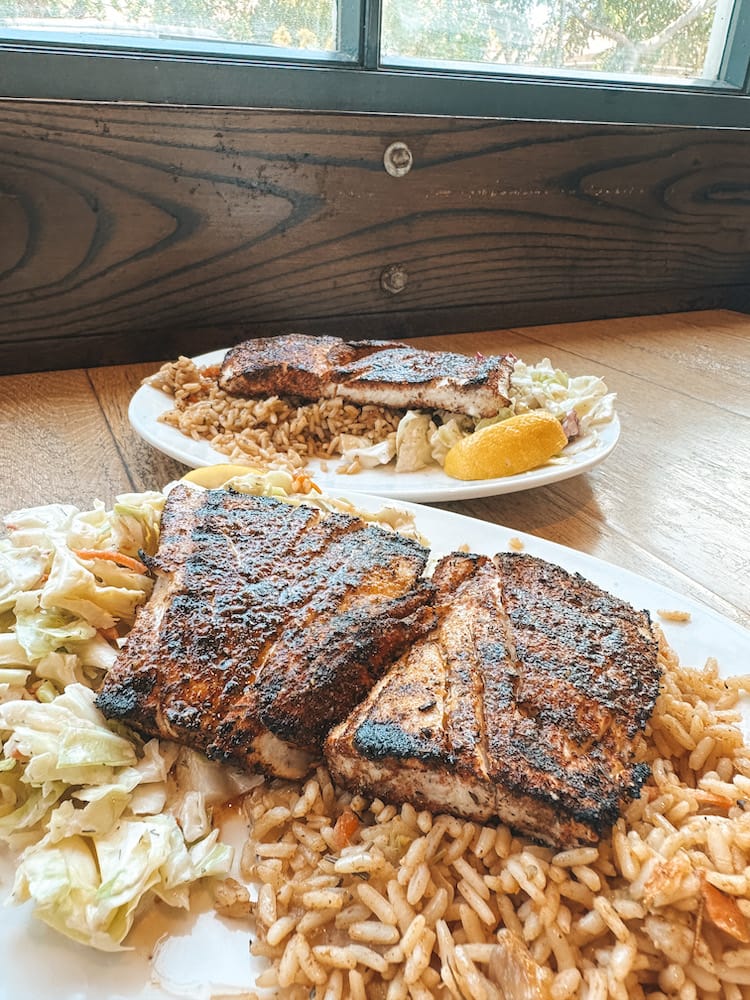 Two plates that each include two filets of blackened mahi mahi, coleslaw, and rice sitting on a wooden table.