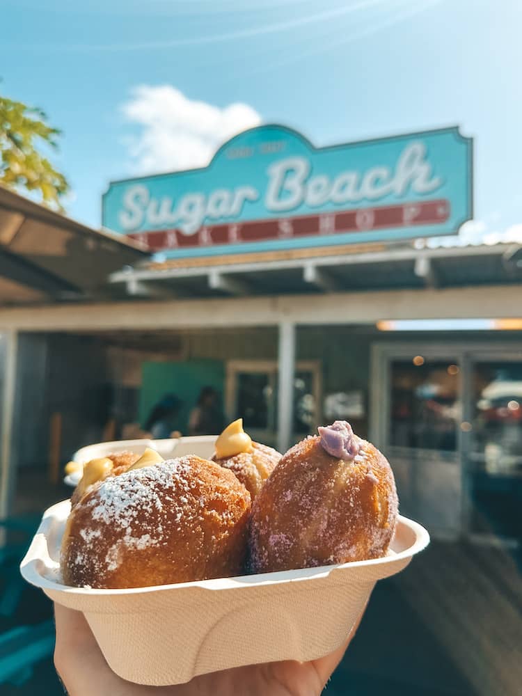 A box of four stuffed malasadas in front of the sign for Sugar Beach Bake Shop, one of the best places to eat in Maui.