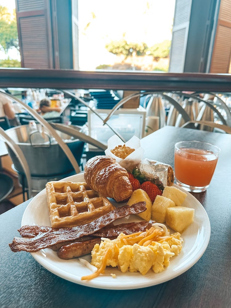 A breakfast spread on a plate with a waffle, croissant, fruit, muffin, breakfast burrito, eggs, bacon, sausage, and juice at a breakfast buffet in Maui.