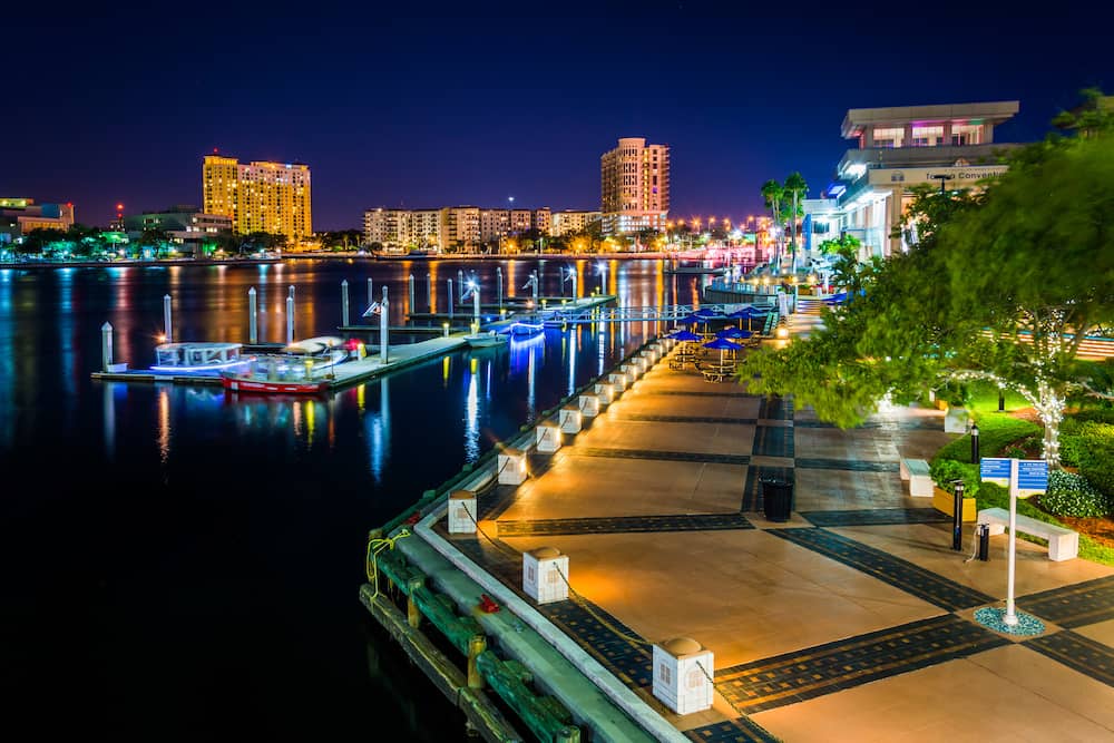 The Tamp Riverwalk at night, with lots of lights illuminating the buildings and reflecting in the body of water below.
