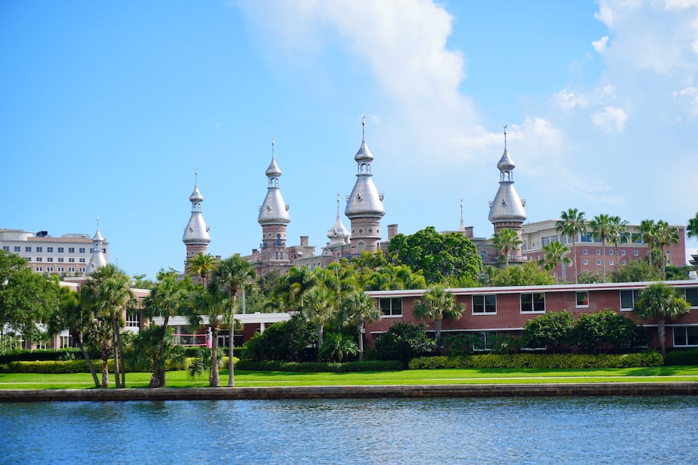 The silver turrets of the University of Florida stick up into the blue sky, attached to a historic red brick building in front of a body of water.