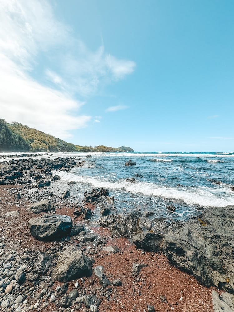 A red sand beach in Maui with black rocks and the blue ocean in the background