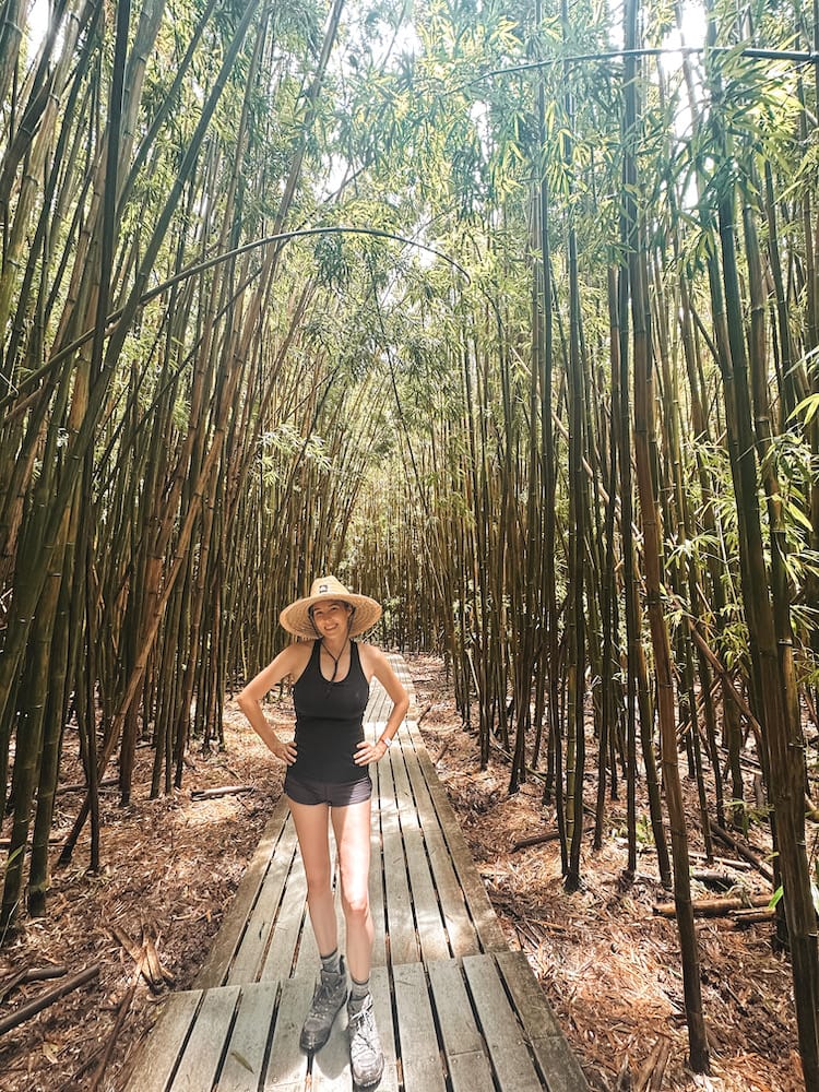 A woman in a black tank top and hiking boots and a straw hat standing in a bamboo forest on the Pipiwai Trailhead in Maui, Hawaii