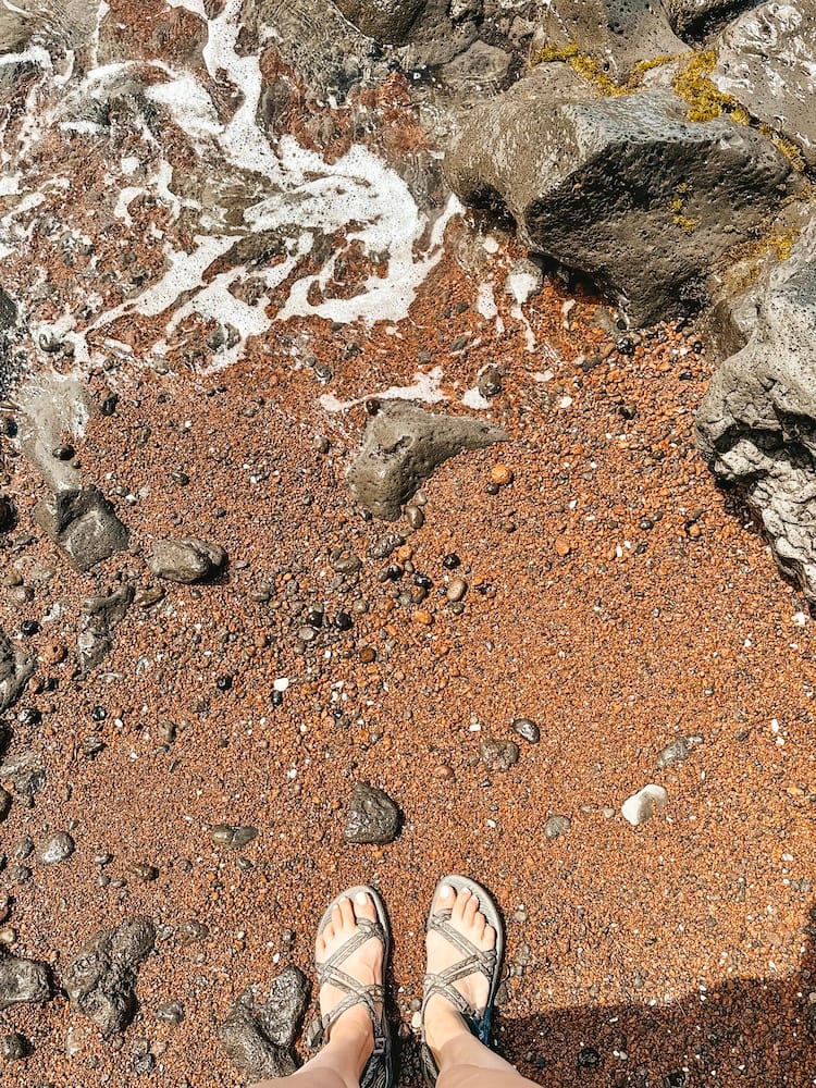 A red sand beach in Maui with black rocks and two feet with sandals standing on the beach
