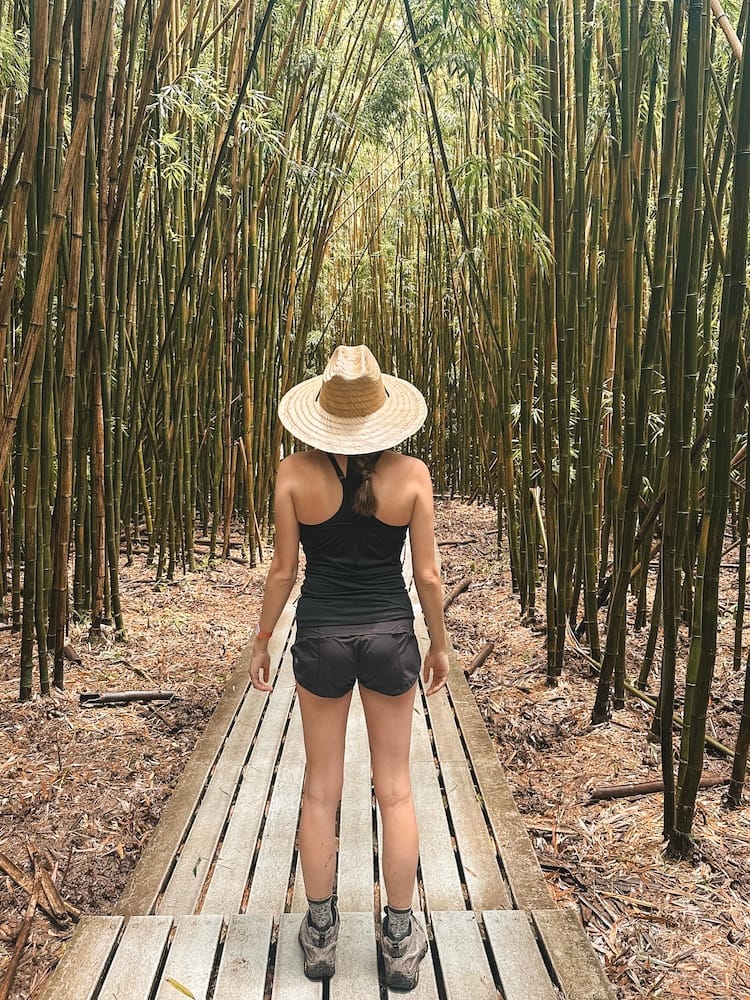 A woman standing in a bamboo forest in Maui wearing hiking boots, black athletic shorts, a black tank top, and a straw hat.