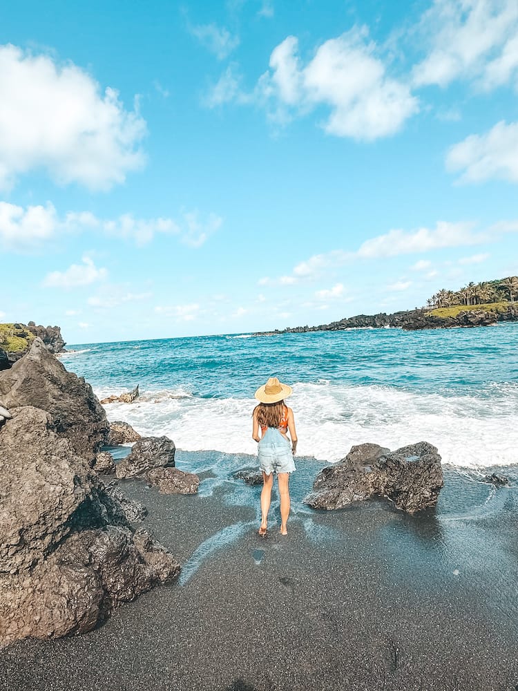 A woman standing on a black sand beach in Maui wearing a straw hat and overalls.