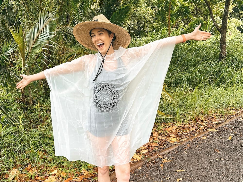 A woman in a straw hat and waterproof poncho standing In front of some green foliage in Hawaii. Preparing for rain is one of the best Road to Hana tips.