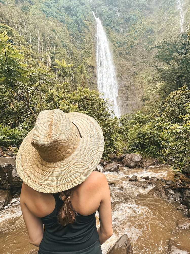 A woman standing in a straw hat and black tank top admiring a waterfall on the Road to Hana