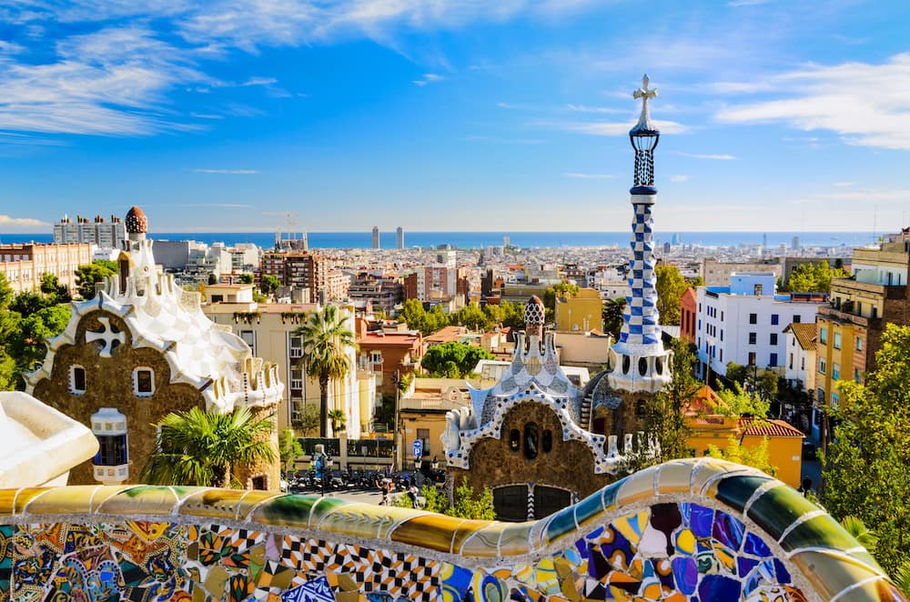 The view from Park Güell in Barcelona, with colorful tiles, unique buildings with white roofs, and several buildings overlooking the ocean.