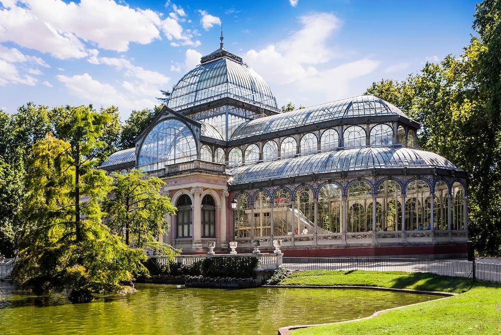 A conservatory with glass ceilings in El Retiro Park in front of a lake and grassy patch in Madrid, one of the best places to visit in spring in Spain.