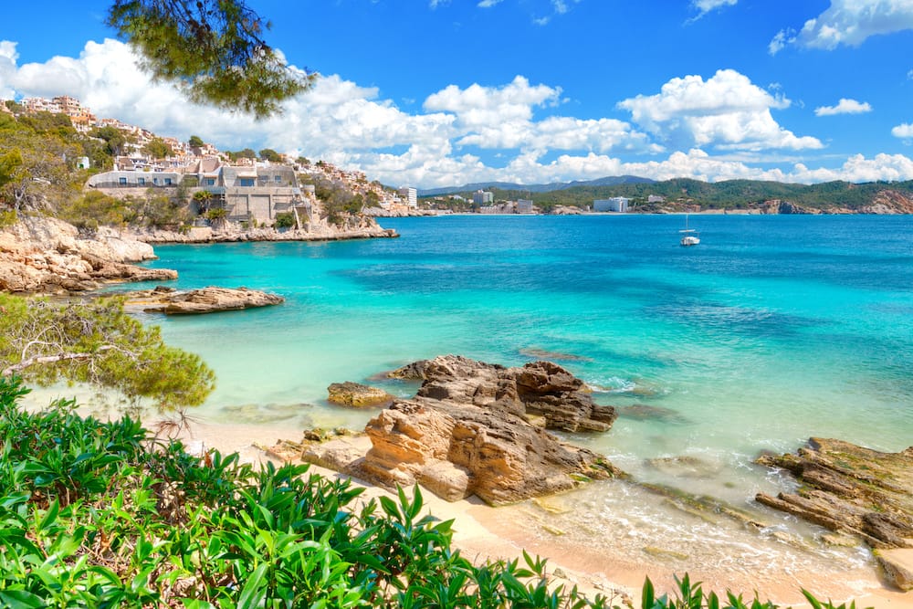 A gorgeous turquoise ocean, fluffy white clouds in a blue sky, rocks on a sandy beach, and greenery in Mallorca, Spain.