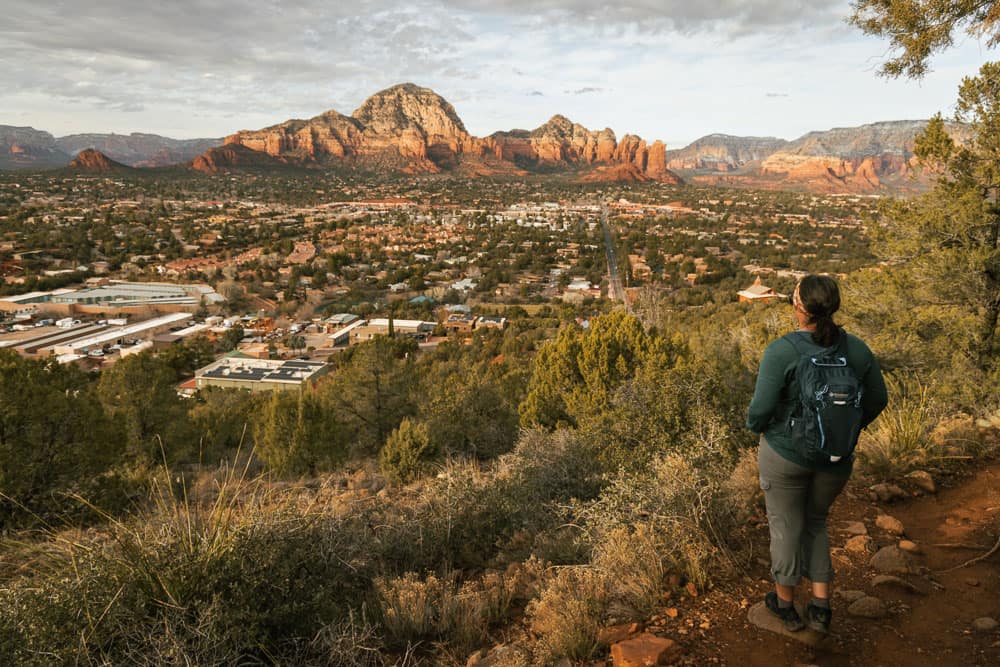 A woman wearing a backpack, pants, and hiking shoes overlooking a scenic overlook in Sedona with the city and red rocks to capture one of the best Instagram spots in Sedona.