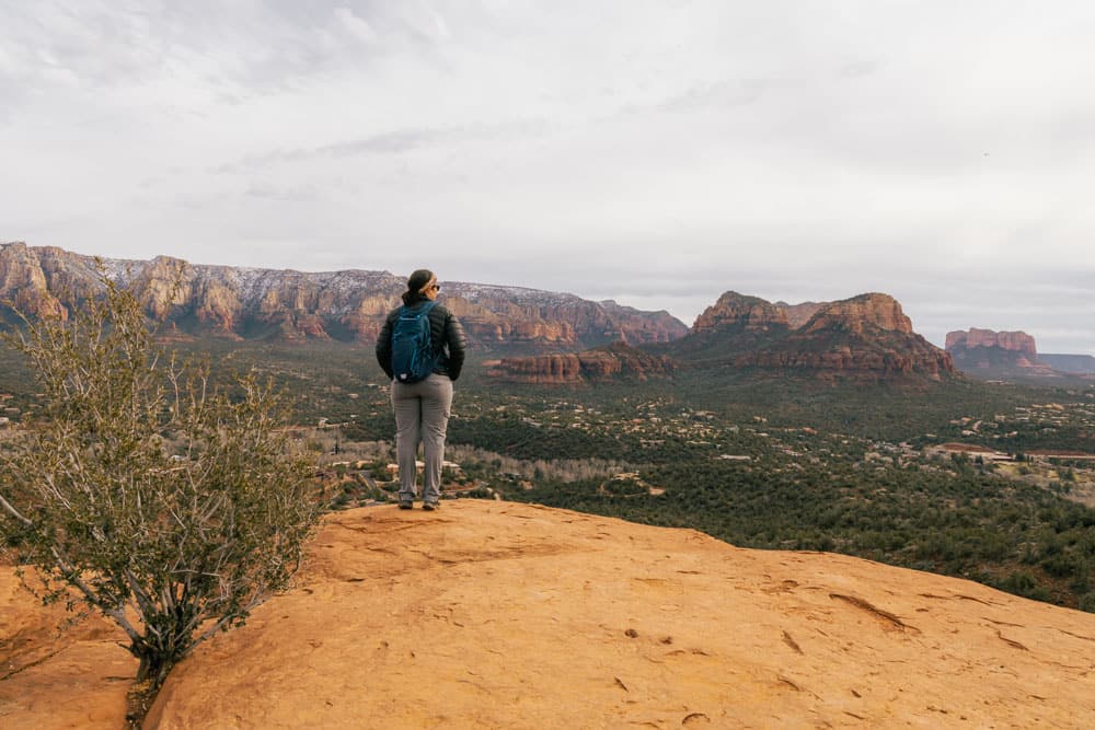 A woman wearing hiking pants, a black puffer jacket, and a blue backpack overlooking the Sedona red rocks and greenery