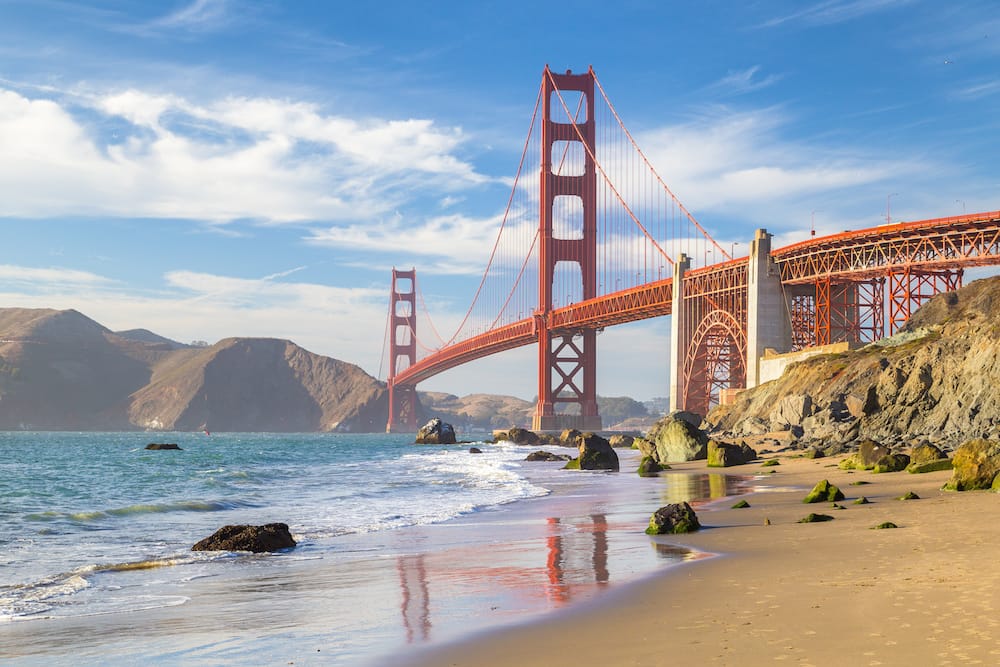 The red Golden Gate Bridge stretches over the sand and water at Baker Beach in San Francisco
