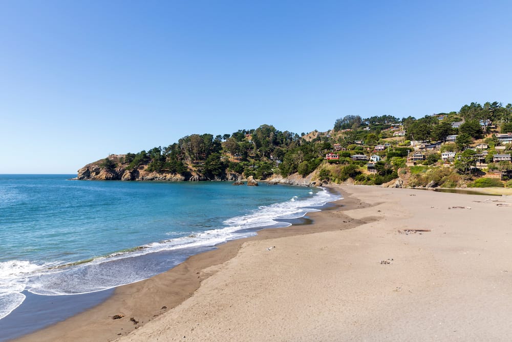 A golden sand beach with bright blue waves and green trees and houses overlooking the beach.