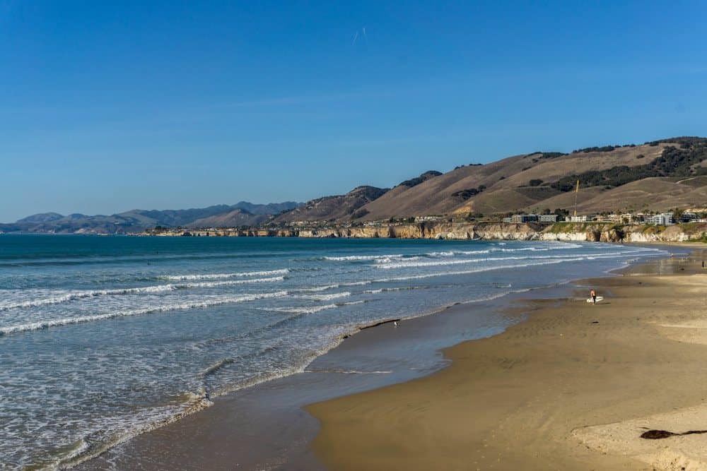 A golden sand beach with rolling hills in the background and blue waves calmly lapping on the shore.